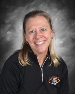 Smiling woman with blonde hair wearing a black zip-up jacket featuring a tiger logo, set against a gray background.