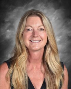 A smiling woman with long, wavy blonde hair, wearing a black top, against a gray backdrop.