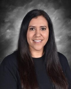 Smiling woman with long black hair, wearing a black shirt, in front of a gray background.