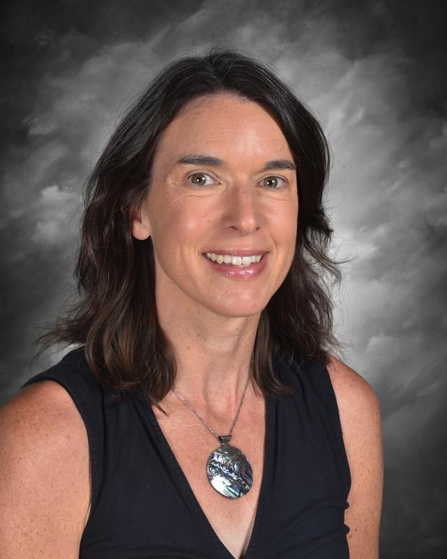 A woman with shoulder-length dark hair smiles, wearing a black top and a decorative pendant against a gray backdrop.