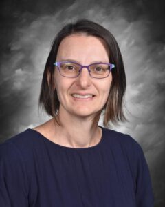 Smiling woman with short brown hair and purple glasses, wearing a navy blue top, against a gray background.