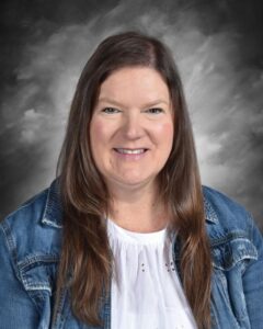 Smiling woman with long brown hair wearing a denim jacket over a white top, set against a gray background.