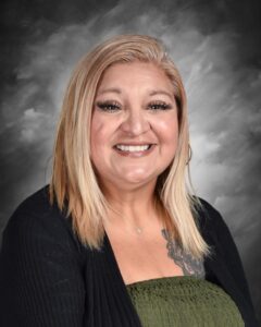 Smiling woman with long blonde hair, wearing a black sweater and green top, set against a soft gray background.