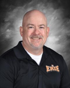 Smiling bald man wearing a black polo shirt with "ERIE" logo, against a gray background.