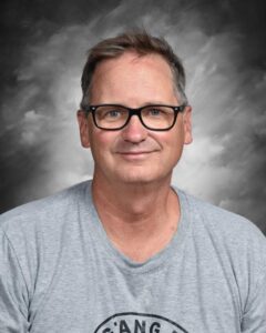 A middle-aged man with glasses smiles, wearing a gray t-shirt, against a softly blurred gray background.