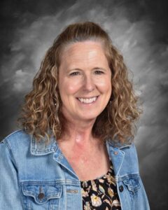 Smiling woman with curly hair, wearing a denim jacket over a floral top, set against a gray background.