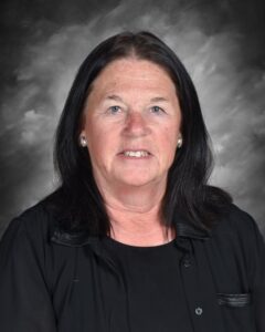 A woman with long black hair smiles, wearing a black top against a gray background.