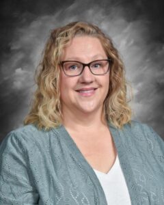 A smiling woman with curly blonde hair, wearing glasses and a light green cardigan, against a gray backdrop.