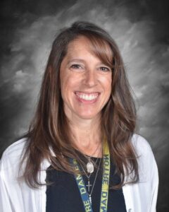 Smiling woman with long brown hair, wearing a black shirt and a lanyard, against a gray background.