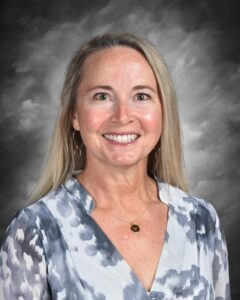 Smiling woman with long blonde hair, wearing a patterned blouse and jewelry, against a gray backdrop.