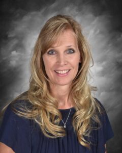 A woman with long, wavy blonde hair smiles in front of a gray backdrop, wearing a dark blue top and a necklace.