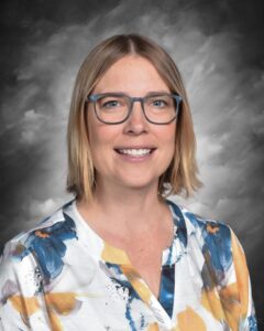 A smiling woman with shoulder-length hair, wearing glasses and a floral shirt, against a gray background.