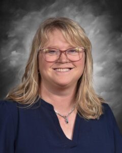 A smiling woman with blonde hair and glasses, wearing a navy top, poses against a gray background.