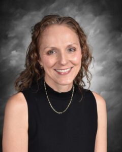 Smiling woman with curly hair, wearing a black top and gold necklace, against a gray background.