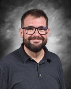 A man with a beard and glasses smiles, wearing a dark polo shirt against a cloudy gray background.