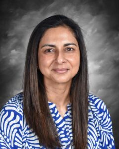 A woman with long black hair smiles, wearing a blue and white patterned blouse against a gray background.
