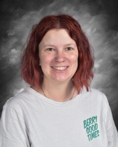 A smiling person with shoulder-length red hair, wearing a gray shirt with green text, poses against a gray backdrop.