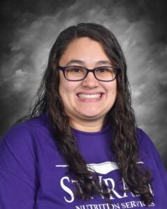 Smiling woman with long, wavy hair wearing a purple shirt with "Nutrition Services" text, set against a gray backdrop.