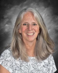 Smiling woman with long gray hair, wearing a leaf-patterned top, set against a gray background.