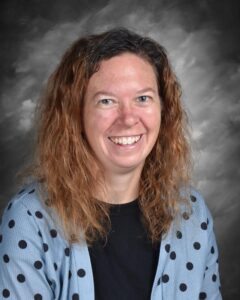 A woman with curly hair smiles, wearing a polka dot cardigan against a gray background.
