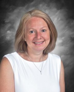Smiling woman with shoulder-length blonde hair, wearing a white top, against a gray background.