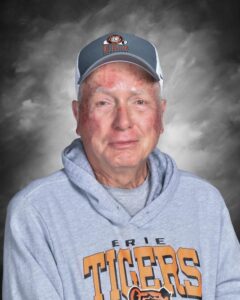 Elderly man with a warm smile, wearing a gray hoodie and a cap, against a soft, cloudy background.