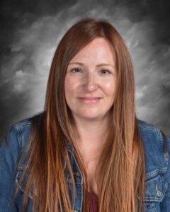 A woman with long, straight brown hair wearing a denim jacket smiles against a gray background.