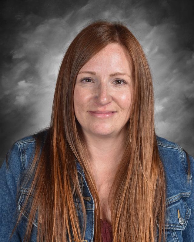 A woman with long, straight brown hair wearing a denim jacket smiles against a gray background.