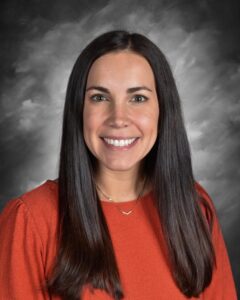 A smiling woman with long dark hair wearing an orange top, set against a gray background.