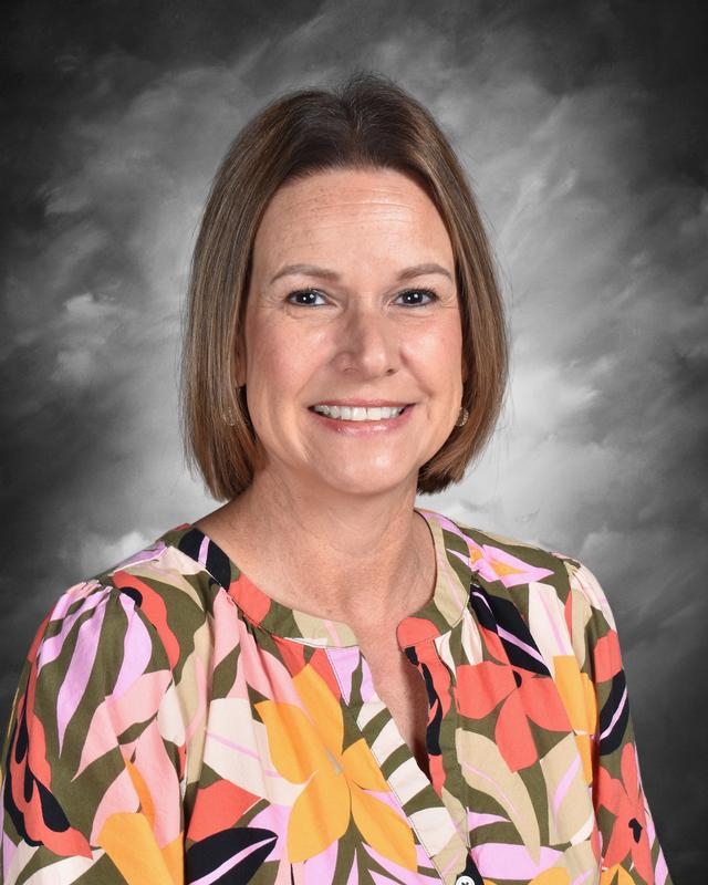Smiling woman with short brown hair wearing a colorful floral blouse, set against a gray background.