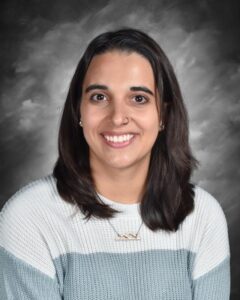 Smiling young woman with shoulder-length dark hair, wearing a light sweater and a mountain necklace, against a gray backdrop.