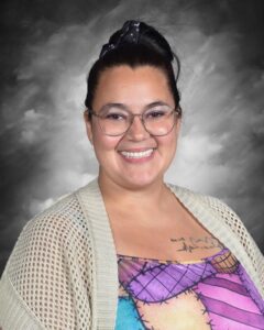 A smiling woman with glasses and a hair bow wears a colorful patterned top and a light sweater against a gray background.