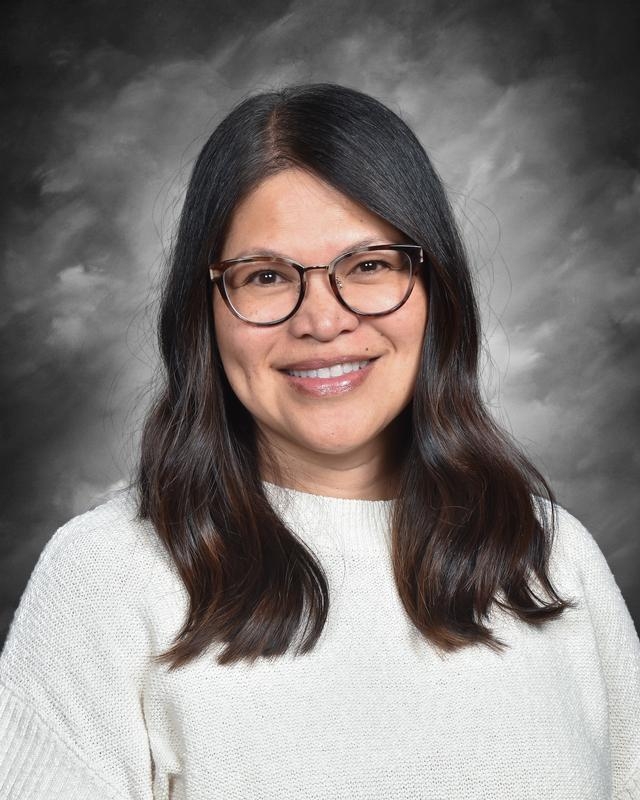 A woman with long, dark hair and glasses smiles warmly, wearing a white sweater against a gray background.