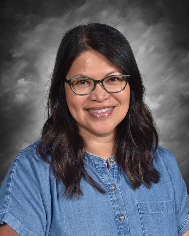 Mujer sonriente con pelo largo y oscuro y gafas, vestida con una camisa vaquera, sobre un fondo gris.