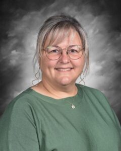 Smiling woman with glasses and light hair, wearing a green top, against a gray background.
