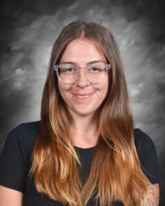 Smiling young woman with long hair and glasses, wearing a black shirt, against a soft gray background.