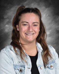 Smiling woman with long brown hair in a denim jacket against a gray background.