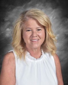 Smiling woman with shoulder-length blonde hair wearing a white blouse, set against a gray background.