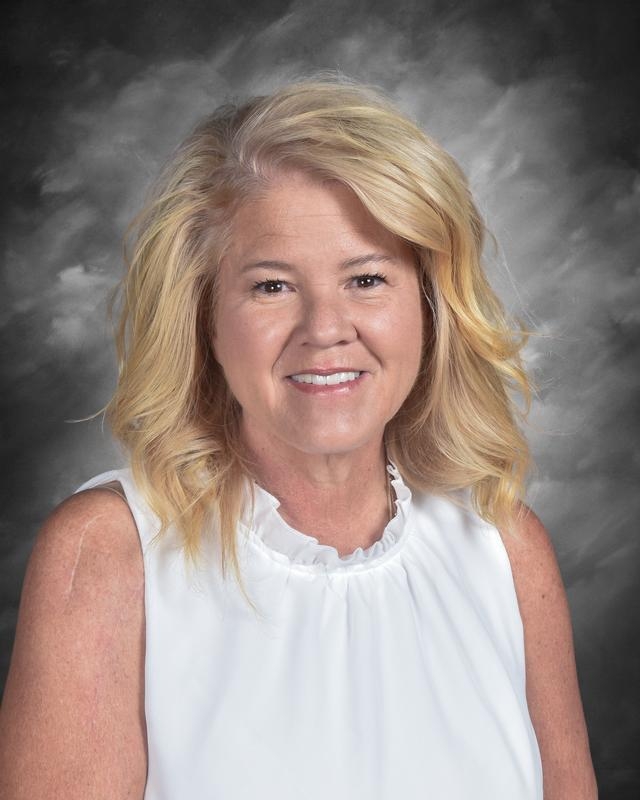 Smiling woman with shoulder-length blonde hair wearing a white blouse, set against a gray background.
