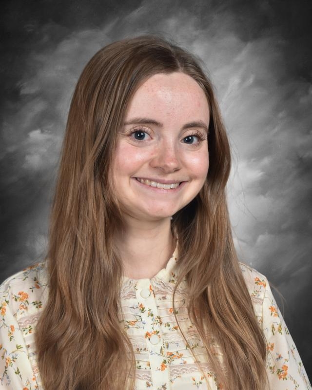 A smiling young woman with long hair wearing a floral blouse, set against a gray background.