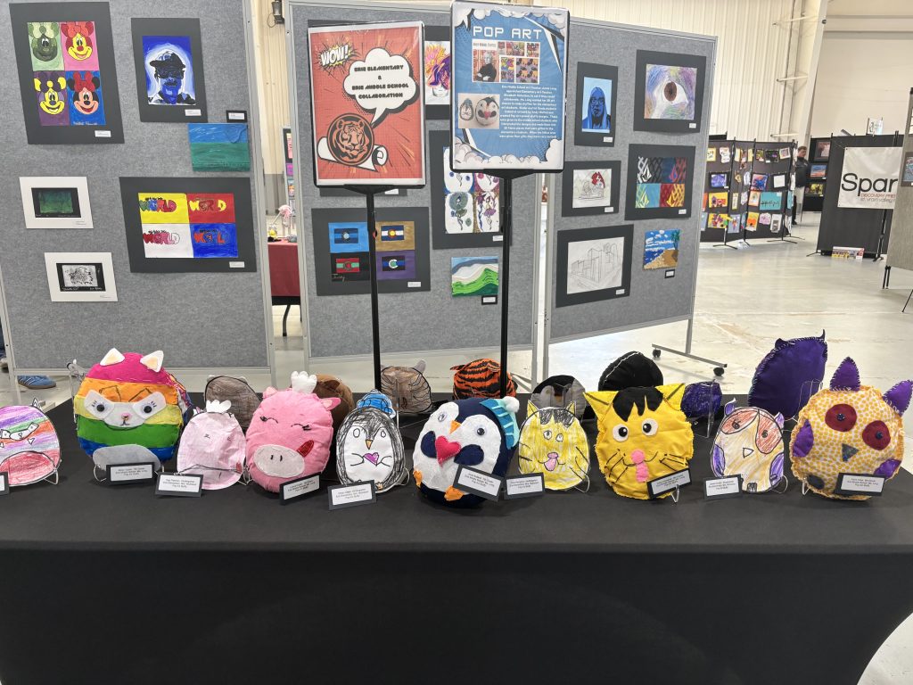 Colorful, handmade plush animal faces displayed on a table, with various artworks in the background at an art show.